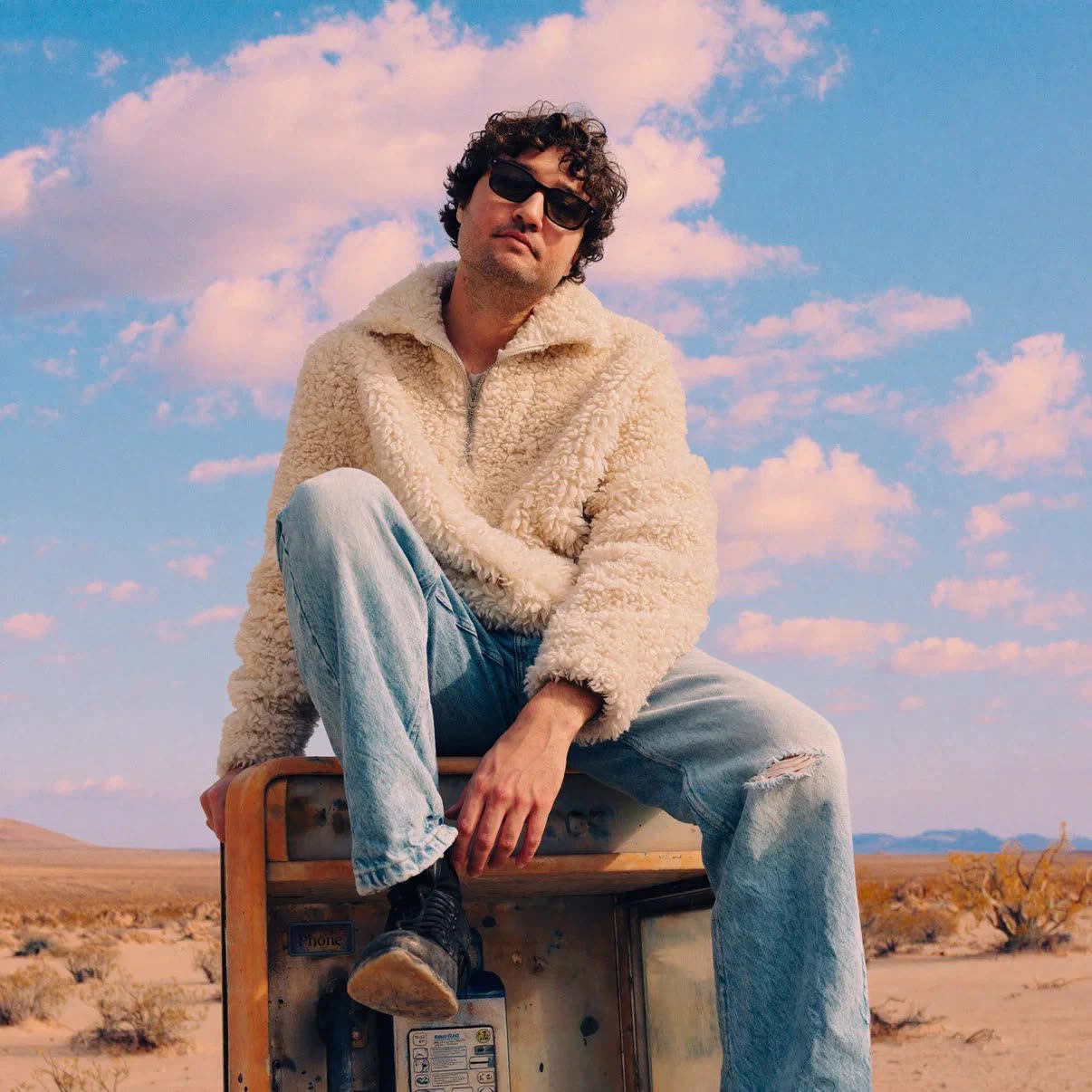 Grynn sitting on an old payphone in the desert under a pink-clouded sky